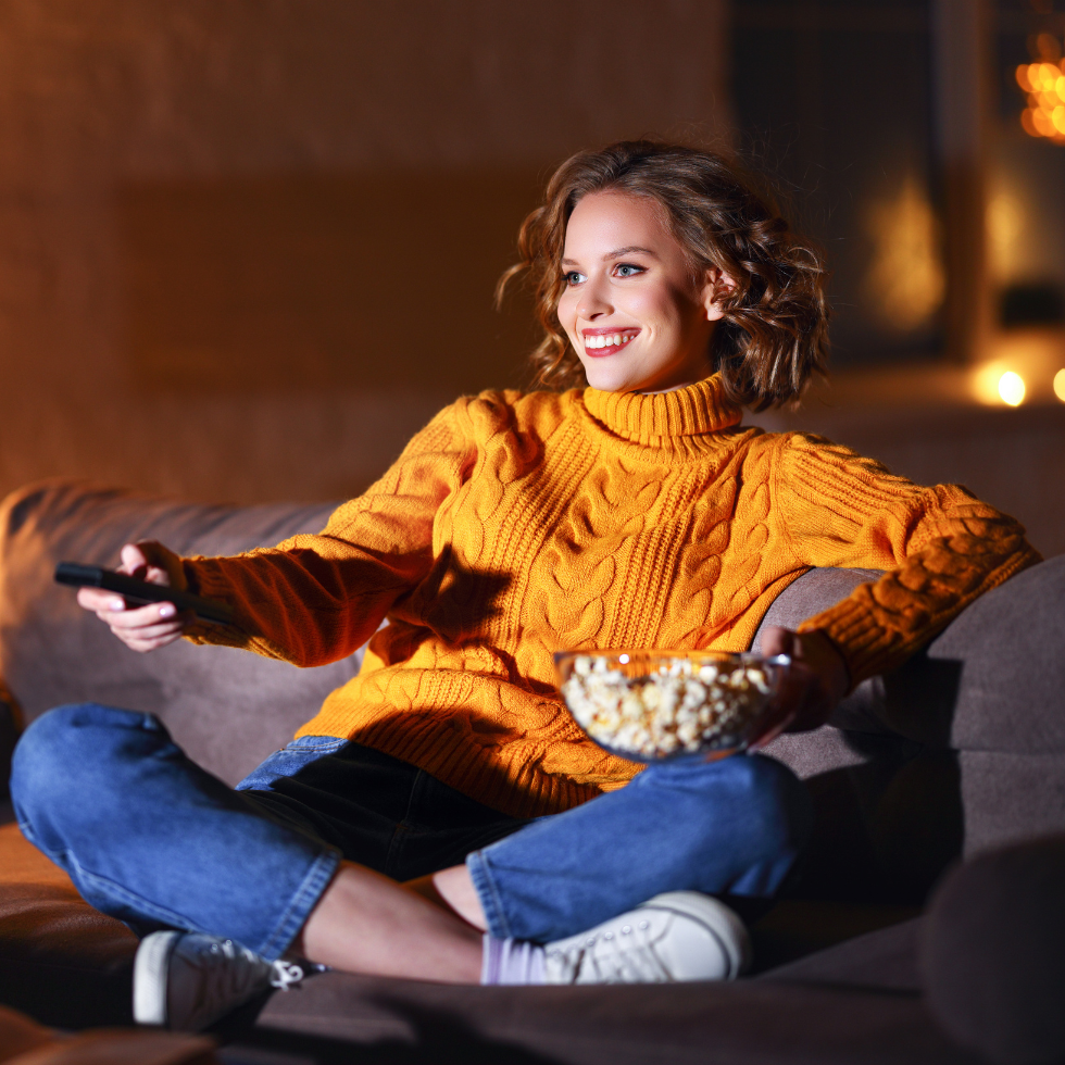 Frau sitzt aus Sofa und schaltet den Fernseher ein während sie eine Schale Popcorn auf dem Schoß hat. Sie möchte Filme und Serien schauen, die perfekt zum Herbst passen.