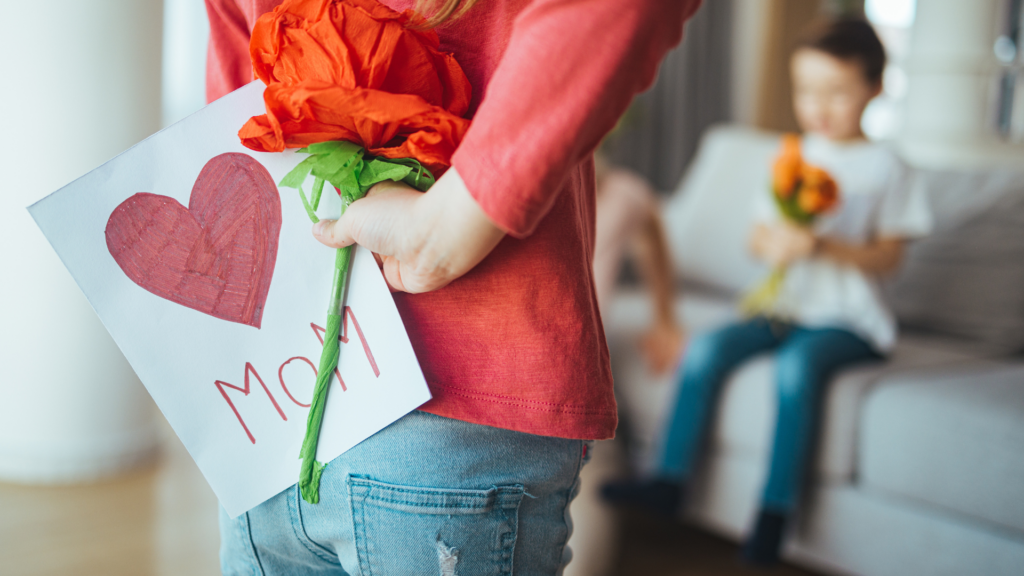 Muttertag: Kind hält hinter dem Rücken eine selbstgemachte Blume und eine Grußkarte für ihre Mutter