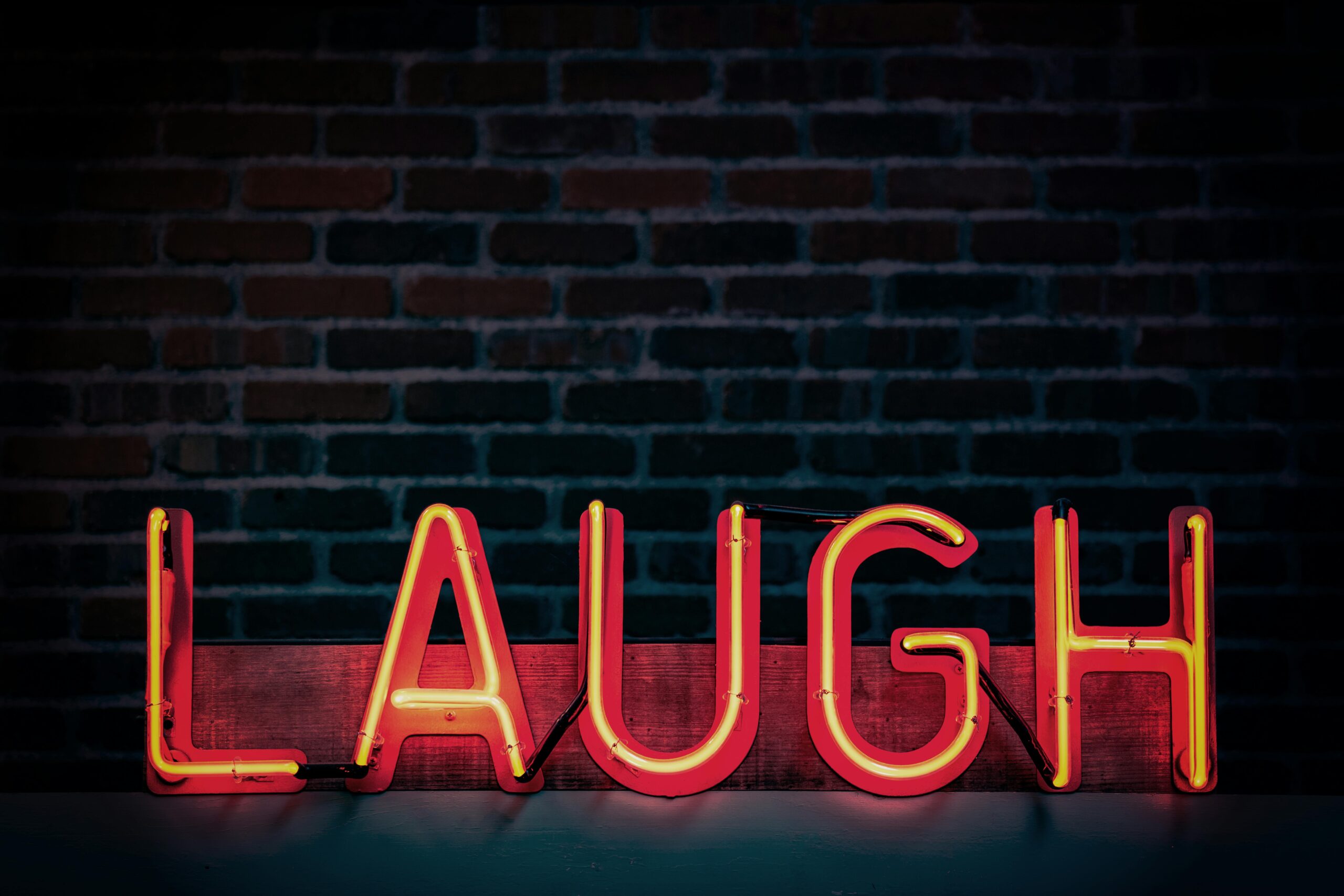 Wie sich Humor auf unsere Psychoe auswirkt: Auf dem Bild ist eine Leuchtschrift mit dem Schriftzug "Laugh" zu sehen, im Hintergrund eine dunkle Ziegelmauer