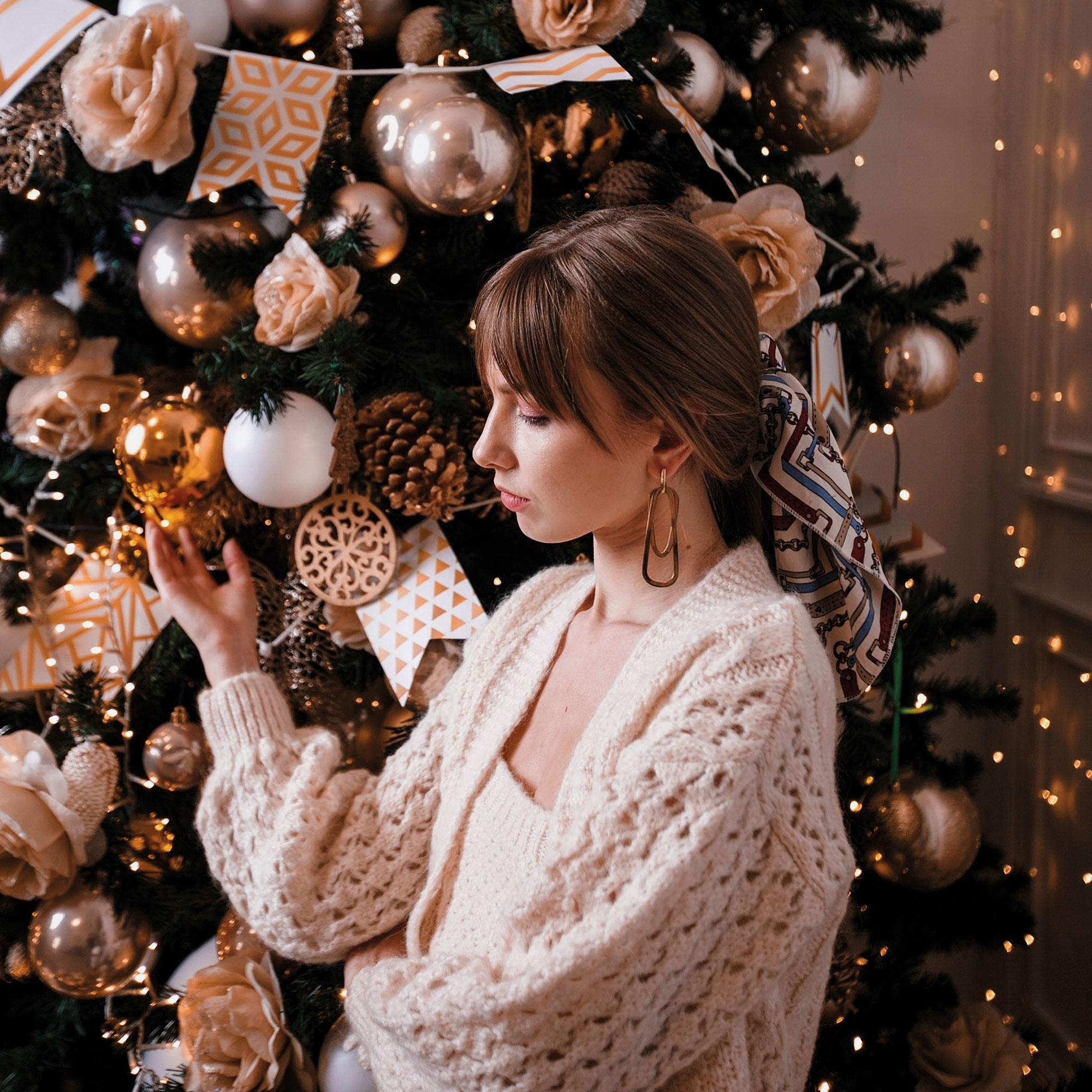 Weihnachtsstress: Frau in beigen Strick-Klamotten steht traurig uneben einem gold geschmückten Christbaum.