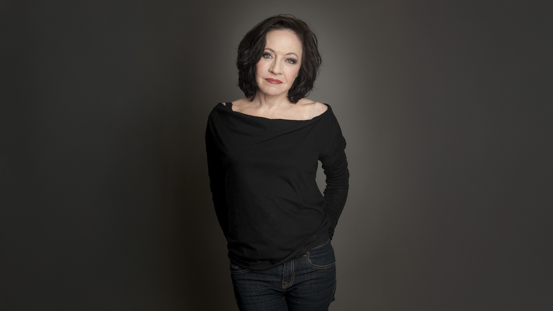 Maria Bill: Ganzkörperporträt einer Frau in schwarzem Oberteil vor neutralem Hintergrund.