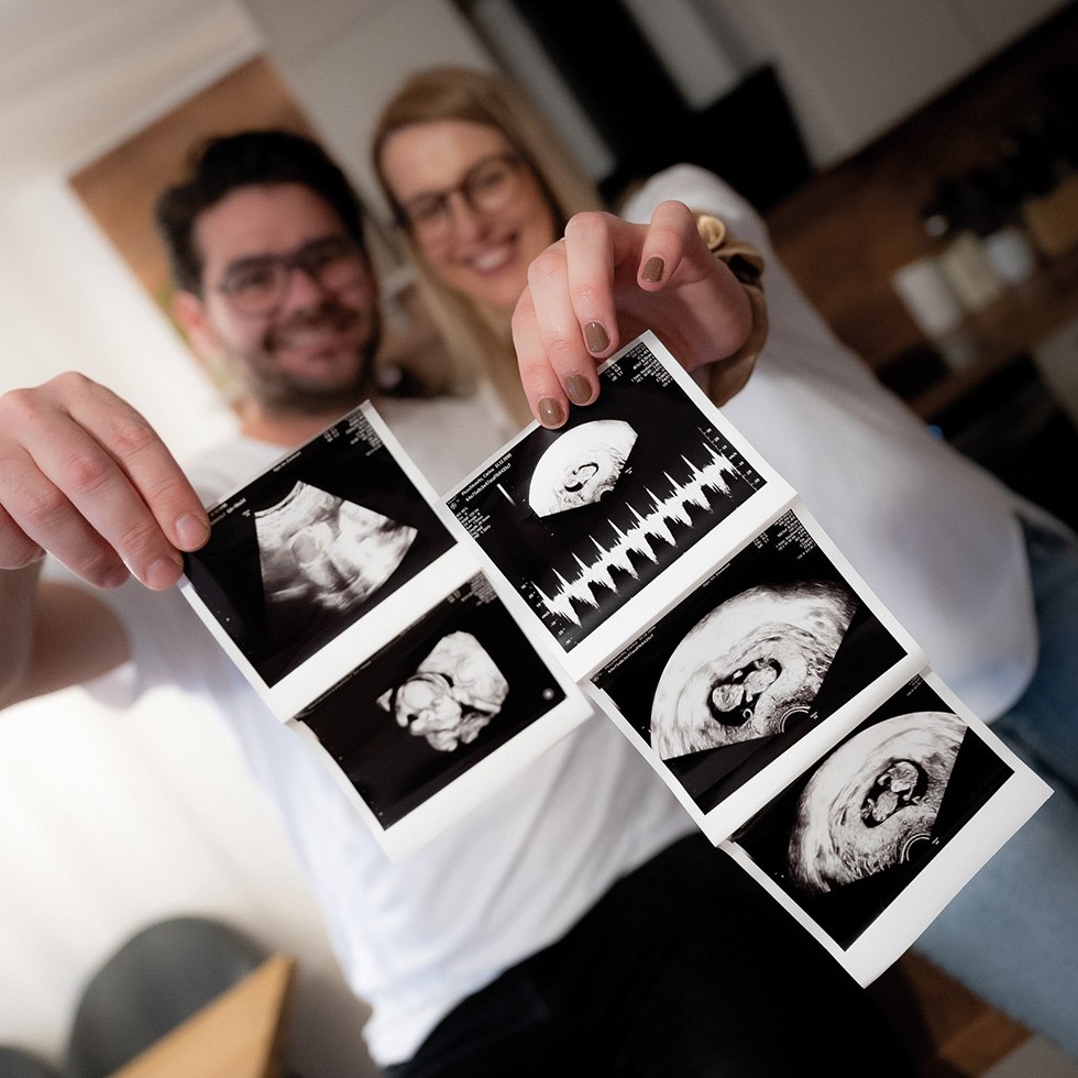 Ein Paar hält Ultraschallbilder in die Kamera und zeigt mehrere Aufnahmen ihres ungeborenen Kindes.